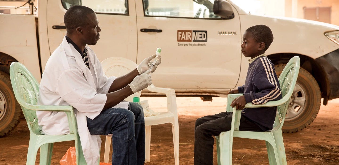 Ein FAIRMED-Mitarbeiter in Kamerun bereitet eine Spritze vor. Er sitzt auf einem weissen Plastikstuhl, im Gegenüber ein kleiner Junge. Sie sitzen im Freien, im Hintergrund ist ein weisses Auto mit einer FAIRMED Beschriftung zu sehen.