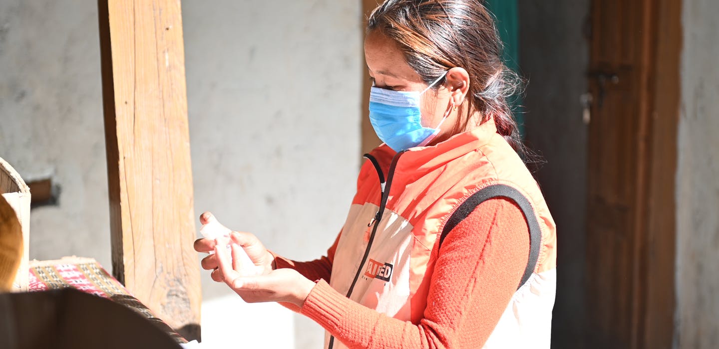 Unsere Gesundheitsmitarbeuterin Gyan, mit Mundschutz und in einem orangen Pullover und einer FAIRMED-Weste, steht vor einem Haus und bereitet eine Behandlung vor.