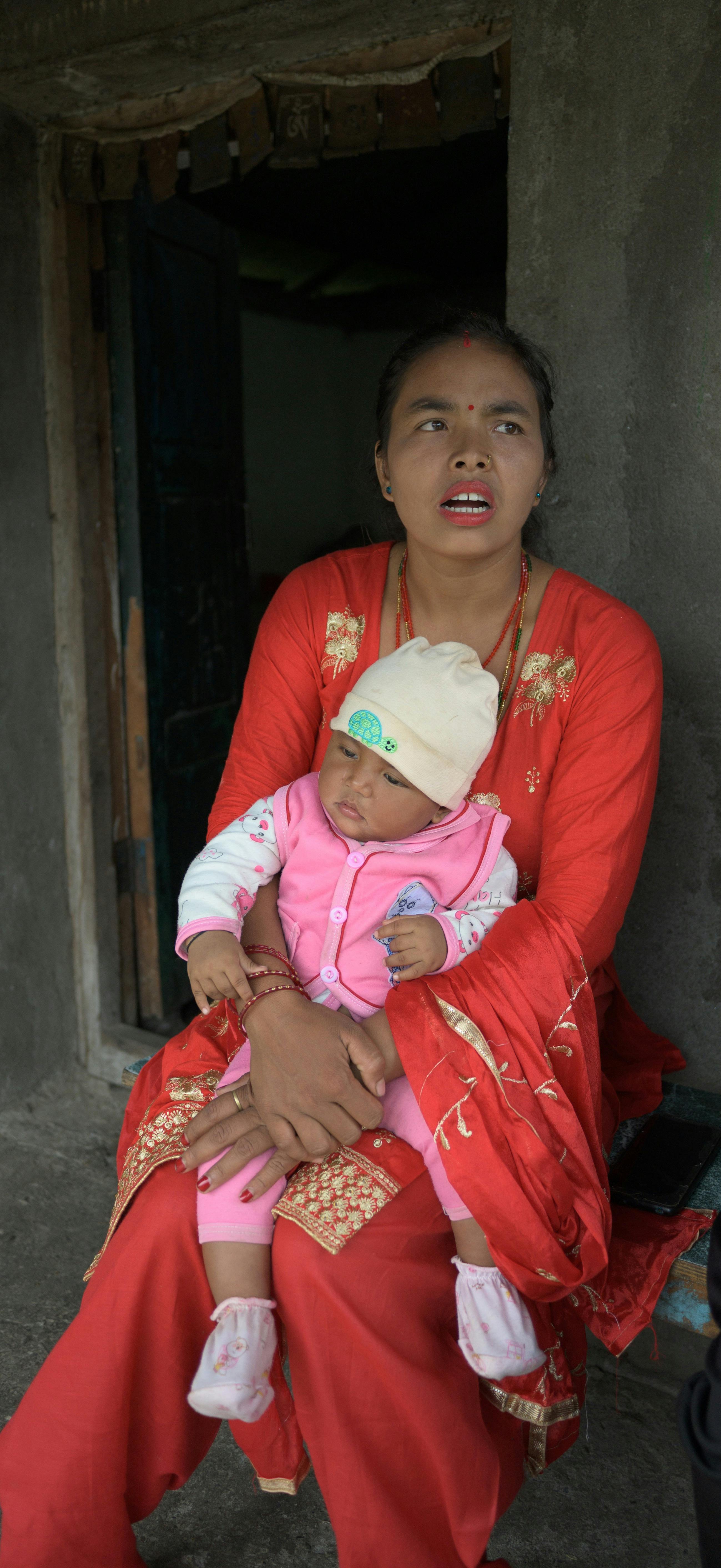 Nani Ram sitzt in einem roten Kleid vor ihrem Haus mit ihrem Kind auf ihrem Schoss.