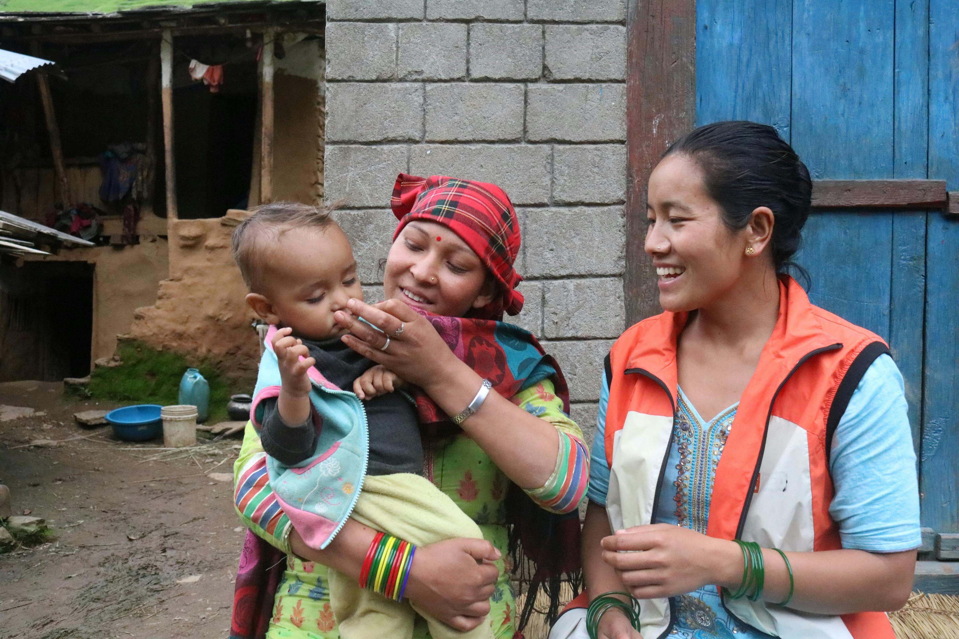 Auf dem Bild ist unsere Gesundheitshelferin Gyan zu sehen. Sie guckt ein Baby an, das auf dem Arm seiner Mutter sitzt, und lächelt das Baby an. Alle drei stehen im Freien vor einer Hütte und sind bunt gekleidet.
