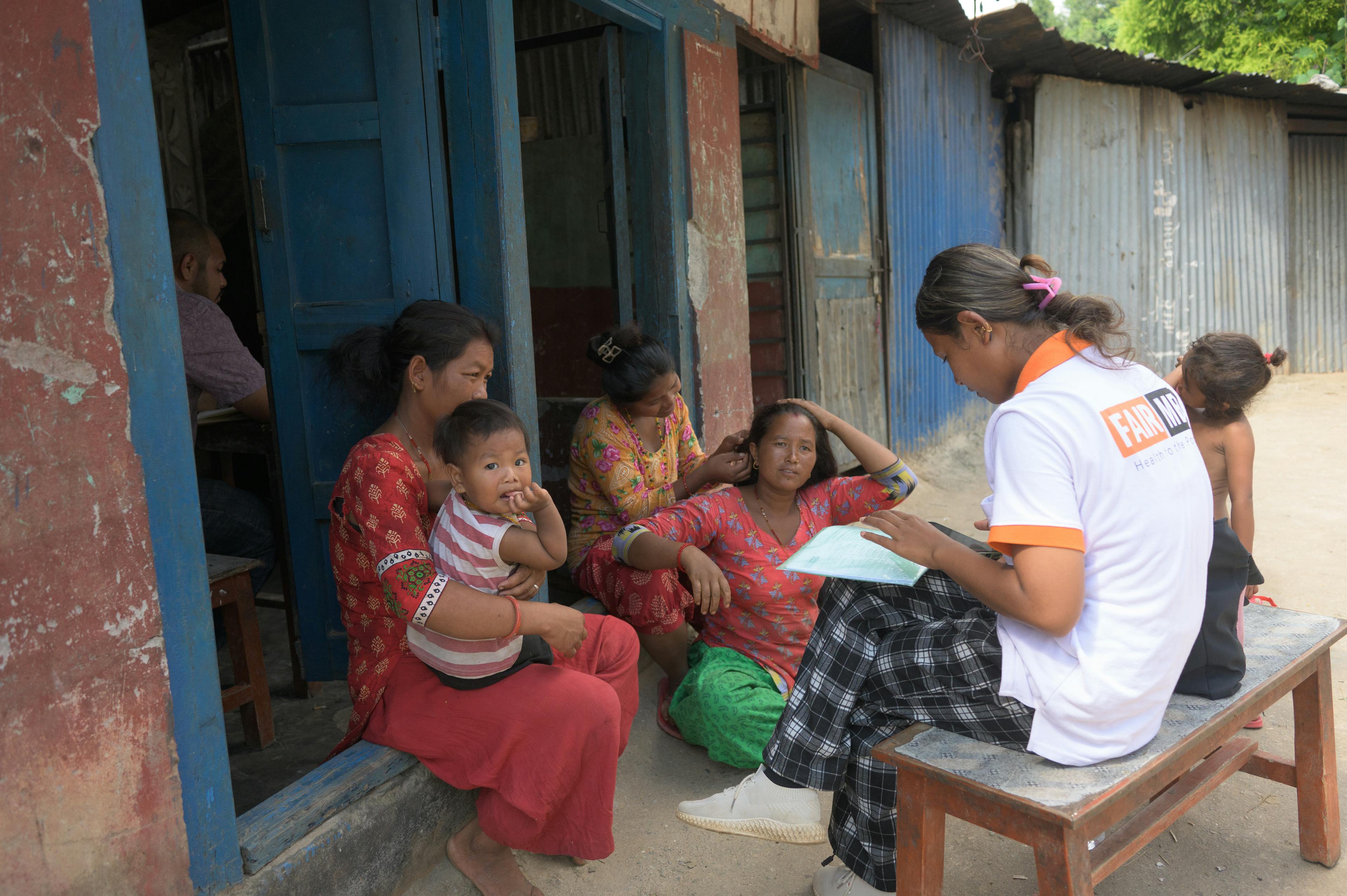 Sujita est assise devant une maison avec trois mères et deux jeunes enfants et les informe sur des sujets de santé.